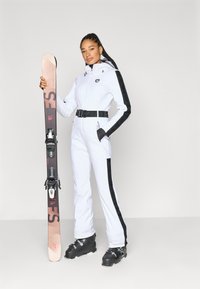 Λευκή στολή σκι με μαύρες λεπτομέρειες, με ζώνη και τσέπες με φερμουάρ. Ένα άτομο κρατά ροζ και μαύρα σκι με δεσίματα, φοράει μαύρα παπούτσια σκι.