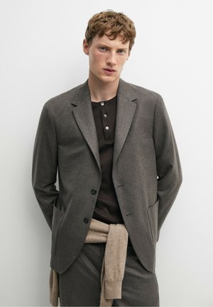 Jeune homme aux cheveux bouclés portant un blazer gris foncé, une chemise noire boutonnée et un pull beige noué autour de la taille, sur fond blanc.