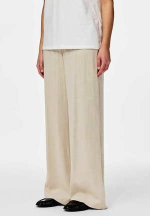 Person som har på seg løse beige linbukser, hvit skjorte og svarte flate sko, står mot en ensfarget hvit bakgrunn.