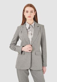 Blazer grigio a righe con revers a punta, chiusura a un bottone e due tasche frontali. Abbinato a una camicia con colletto grigia chiara.
