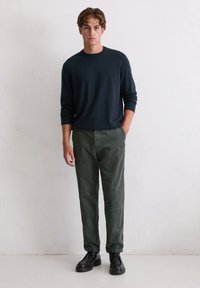 Pull en jersey à col rond bleu marine à manches longues, associé à un pantalon en velours côtelé vert à rayures verticales ; porté avec des chaussures noires à lacets.