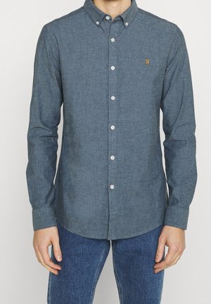 Homme portant une chemise chambray grise à manches longues boutonnée avec un petit logo jaune brodé sur la poitrine et un jean bleu, debout sur fond blanc.