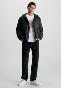 Giacca nera con zip e cappuccio, polsini elastici. Maglietta bianca con stampa grafica beige, pantaloni neri e sneakers bianche. Design semplice e informale.