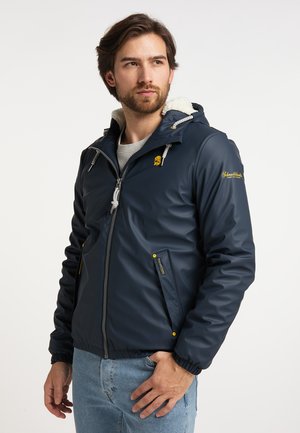 Mann trägt eine dunkelblaue Kapuzenjacke mit Fleecefutter und Reißverschluss sowie hellblaue Jeans und schaut leicht zur Seite.