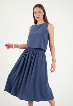 Mujer con top azul sin mangas y falda ancha a juego posando sobre fondo blanco, lleva pendientes largos, pulsera y reloj.