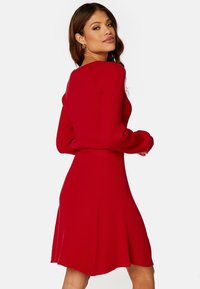 Röd ribbad stickad klänning med långa ärmar, figurnära bodice och utsvängd kjol. Tyget har en strukturerad yta och en väldefinierad midja.