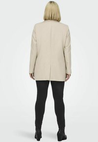 Beige sako s rovnými liniemi, dlouhými rukávy a jedním rozparkem na zadní straně, nošeno přes černé legíny a kotníkové boty. Hladká textura.