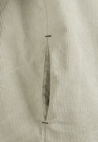 Gros plan sur un tissu en velours côtelé beige avec une poche fendue verticale délimitée par des coutures surpiquées et des renforts en point de barrette en haut et en bas.