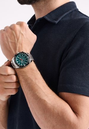 Sølv rustfrit stål ur med grøn skive, tre under-skiver og røde detaljer, båret på et håndled, sammen med en textured navy polo skjorte.