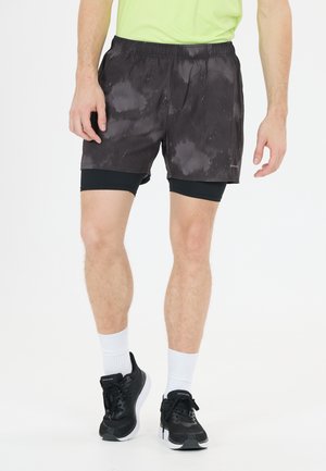 Man die zwarte sportshorts met patroon draagt, met zwarte compressietights eronder, witte sokken en zwarte hardloopschoenen.