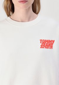 Bílá mikina s žebrovaným výstřihem a výraznou červenou grafikou s nápisem "TOMMY JEANS NEW YORK CITY" na levé části hrudi.