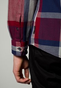 Vänster arm och hand av en person som bär en röd, blå och vit rutiga skjorta med mörka knappar och svarta byxor mot en enfärgad bakgrund.