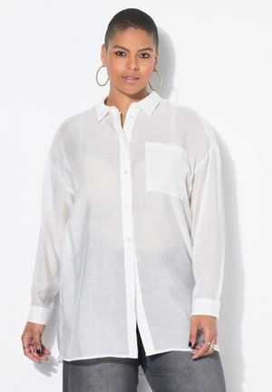 Kvinde med kort hår iført store store øreringe, en transparent hvid skjorte med knapper og grå bukser, stående mod en hvid baggrund.