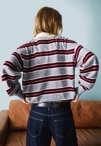 Bombažna jopica v sivi barvi z rdečimi in belimi vodoravnimi črtami. Ohlapen kroj, spuščena ramena, v kombinaciji s temnimi kavbojkami in rjavim pasom.