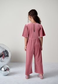 Combinaison en velours côtelé rose avec manches ballon courtes, taille élastique et une ouverture en forme de goutte dans le dos. Associée à des chaussures colorées sans lacets.