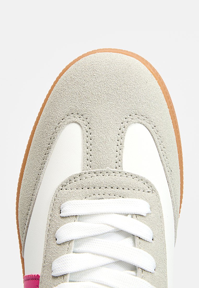 Zapatilla con parte superior de ante gris, acentos de cuero blanco y suela de goma texturizada. Presenta cordones blancos y un detalle en rosa en el costado.