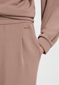 Lichtbruine sportieve broek met een gladde textuur, voorzien van een zijzak en een klein gouden "BOSS" logo op de tailleband.