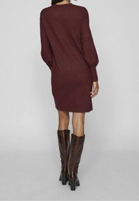 Burgundské pletené šaty se dlouhými nafouknutými rukávy, kulatým výstřihem a délkou ke kolenům, doplněné vysokými hnědými koženými botami.