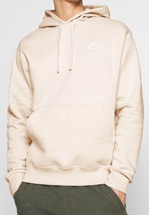 Personne portant un sweat à capuche Nike beige clair avec une poche avant et des cordons, associé à un pantalon vert foncé, les mains dans les poches.