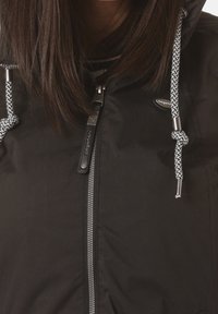 Närbild av en person som bär en svart jacka med dragkedja och svart-vita flätade snoddar på huvan.