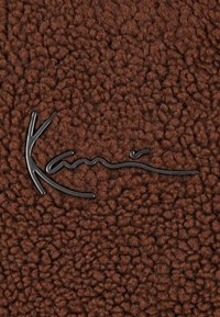 Tessuto marrone con una trama morbida e pelosa, con un logo in metallo nero che assomiglia a una firma stilizzata.