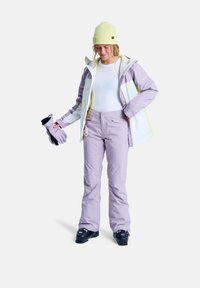 Schneebekleidung mit einer lila und weißen Jacke, dazu passenden lila Hosen und einer hellgelben Mütze. Handschuhe in der Hand mit strukturierten Akzenten.