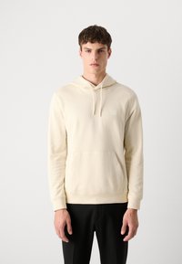 Felpa color crema con tasca a marsupio, cappuccio regolabile con cordino, polsini a coste e un piccolo patch con logo sul davanti. Tessuto morbido.