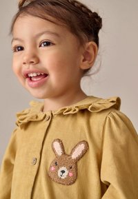 Cămașă din corduroy muștar cu guler volan, având un applique de iepure brun pe piept. Materialul are un design texturat, cu dungi.