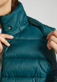 Teal puffjakke med hög krage, ficka med dragkedja på sidan och sydda detaljer. "Pepe Jeans"-logotyp synlig på framsidan.