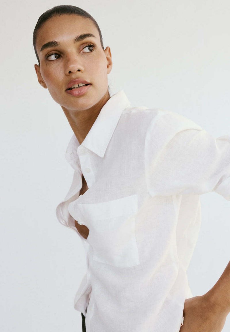 Femme aux cheveux tirés en arrière portant une chemise blanche boutonnée, regardant sur le côté devant un fond clair uni.