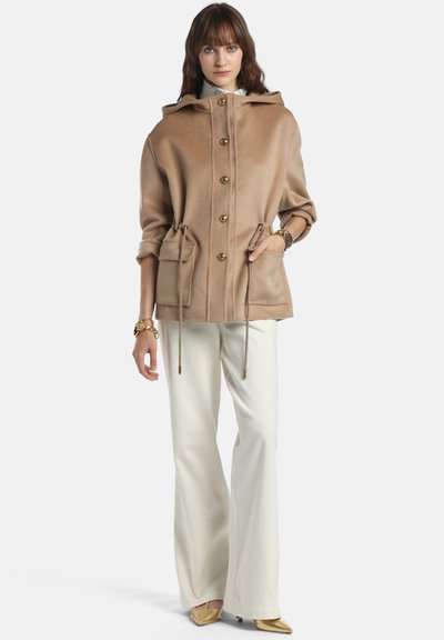 Jachetă din amestec de lână de culoare maro deschis, cu glugă, nasturi aurii, talie cu șnur și două buzunare frontale. Prezintă o croială lejeră și mâneci lungi.