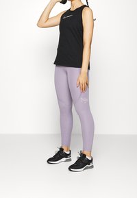 Top negro sin mangas con un logo plateado de Nike, combinado con leggings morados claros que presentan un logo de Nike, y zapatillas deportivas negras con detalles en verde.