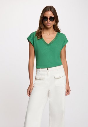 Mujer con gafas de sol oscuras, top verde de manga corta con escote en V y pantalones blancos de talle alto con bolsillos delanteros con solapa, de pie con la mano al lado.
