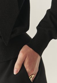 Mână cu inel curbat din aur parțial în buzunarul de la pantaloni negri, purtând un top lung cu mâneci negre, cu manșete și talie ribbate.