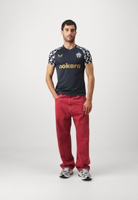Marineblaues Sporttrikot mit einem Schachbrettmuster an den Schultern, goldenem "nokera"-Logo, kombiniert mit roten Jeans und weißen Sneakers. Einfache Hintergrund.