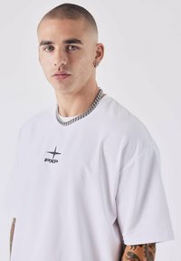 Witte t-shirt met korte mouwen met zwart "PXP" logo en sterontwerp op de borst. Bevat een zilveren ketting en zichtbare tatoeages.