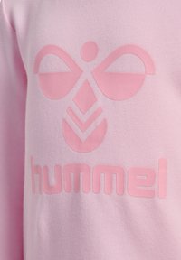 Jasnoróżowy sweter z teksturowanym logo, które przedstawia symetryczne kształty oraz słowo "hummel" wydrukowane wyraźną, zaokrągloną czcionką.