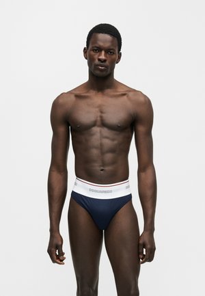 Fit tmavá pleť muža stojí, oblečený v námornícky modrých slipoch s bielym pásom a značkou "DSQUARED2" na jednoduchom bielom pozadí.