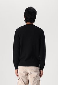 Person steht mit dem Rücken zur Kamera und trägt einen schwarzen langärmeligen Pullover sowie beige Cord-Hose gegen einen schlichten Hintergrund.