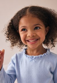 Hellblaues Langarmhemd mit gerafftem Kragen und einer kleinen blauen Schleife als Akzent. Weiche Textur mit einem zarten Muster überall.