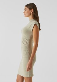 Testhez simuló, rövid ujjú ruha világos bézs színű, sima textúrájú anyagból, oldalsó ráncolt részletekkel, elegáns vonalvezetést bemutatva.