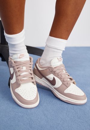 Osoba nosząca białe skarpetki i beżowe buty sportowe Nike z brązowym logo na jasnoniebieskim, wykładzinowym podłożu.