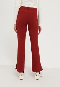 Pantalons flared rouges en tissu extensible, dotés de poches arrière et d'une fente discrète à l'ourlet. Portés avec un haut blanc et des baskets grises.