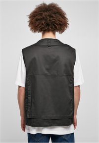 Musta liivi, jossa on sileä pinta, takatasku ja kaulan kohdalla lenkkitiedetaili, puettuna valkoisen lyhythihainen t-paidan päälle.