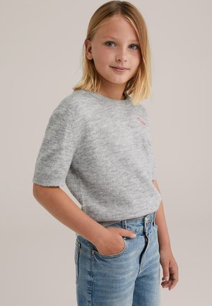 Blond tjej med blå ögon som bär en grå kortärmad tröja och blå jeans, stående med en hand i fickan mot en enfärgad bakgrund.