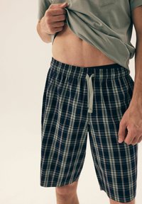 Męskie szorty w kratę charakteryzują się granatowo-zieloną kolorystyką z cienkimi białymi liniami, elastycznym pasem oraz detalem w postaci sznurka.