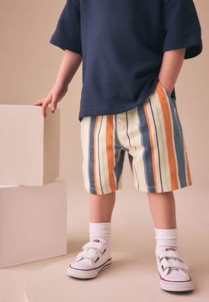 Kind trägt gestreifte Shorts, weiße Sneakers und ein dunkelblaues Hemd, steht neben weißen rechteckigen Blöcken.