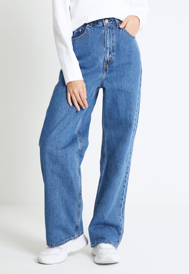 Personne portant un jean bleu ample à taille haute, des baskets blanches et un haut blanc à manches longues, debout avec une main dans la poche.