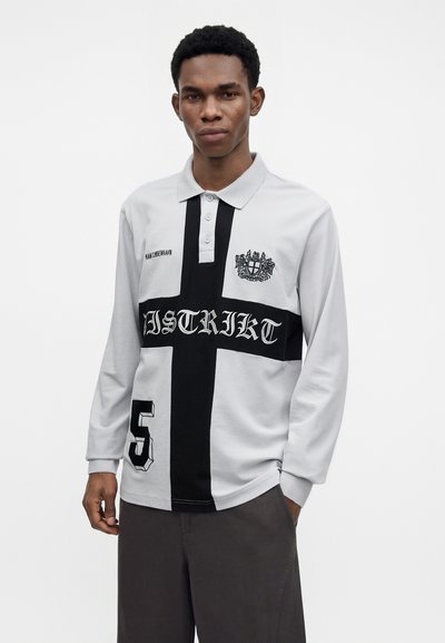 Jeune homme portant un polo blanc à manches longues avec une croix noire, un blason, le texte "DISTRIKT" et le numéro 5, debout sur un fond blanc.