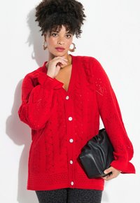 Roter Strickcardigan mit V-Ausschnitt, großen Knöpfen und strukturierten Mustern. Kombiniert mit einer schwarzen Clutch und Leggings im Leopardenmuster.